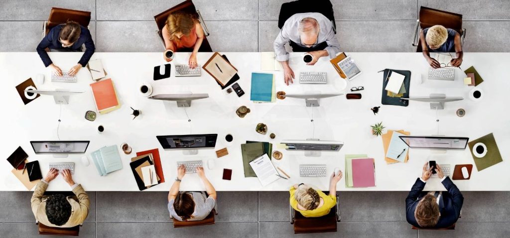 8 people sit together in a long white desk , 4 people each side working for technology company