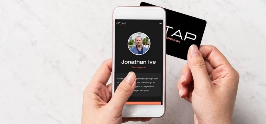 girl's finger is holding smartphone that shown her profile and her left hand is holding a smart business card from gotap on the white desk