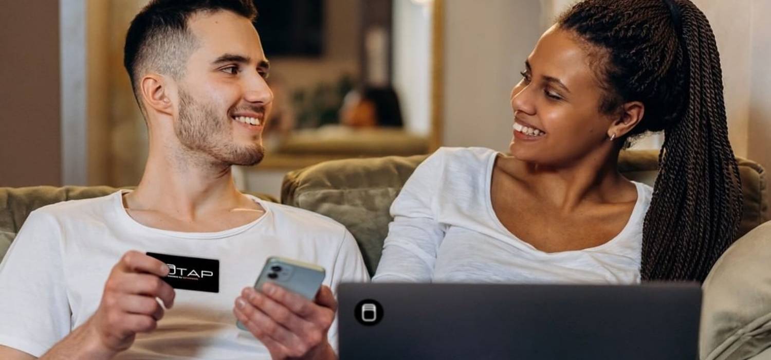 white man and black woman both wear white t-shirt sit together in the sofa while the man show his smart business card to her, and laptop in front of her with smart tag stick on it