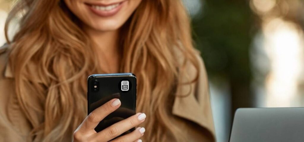 seorang wanita cantik berambut pirang yang cantik menggunakan fungsi NFC di ponselnya dengan tag/stiker pintar GOTP di sisi belakang smartphone