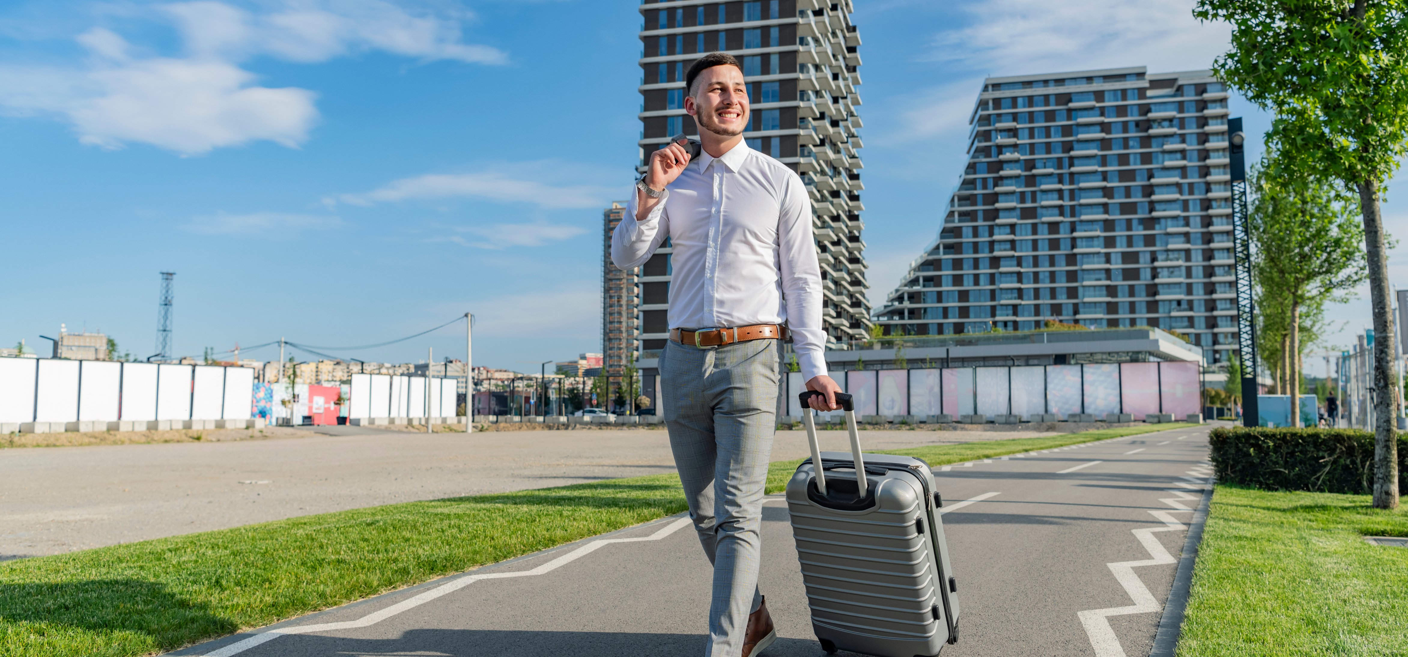 a businessman in a white shirt brown belt and gray pants is walking out of the airport carrying a grey suitcase while smiling