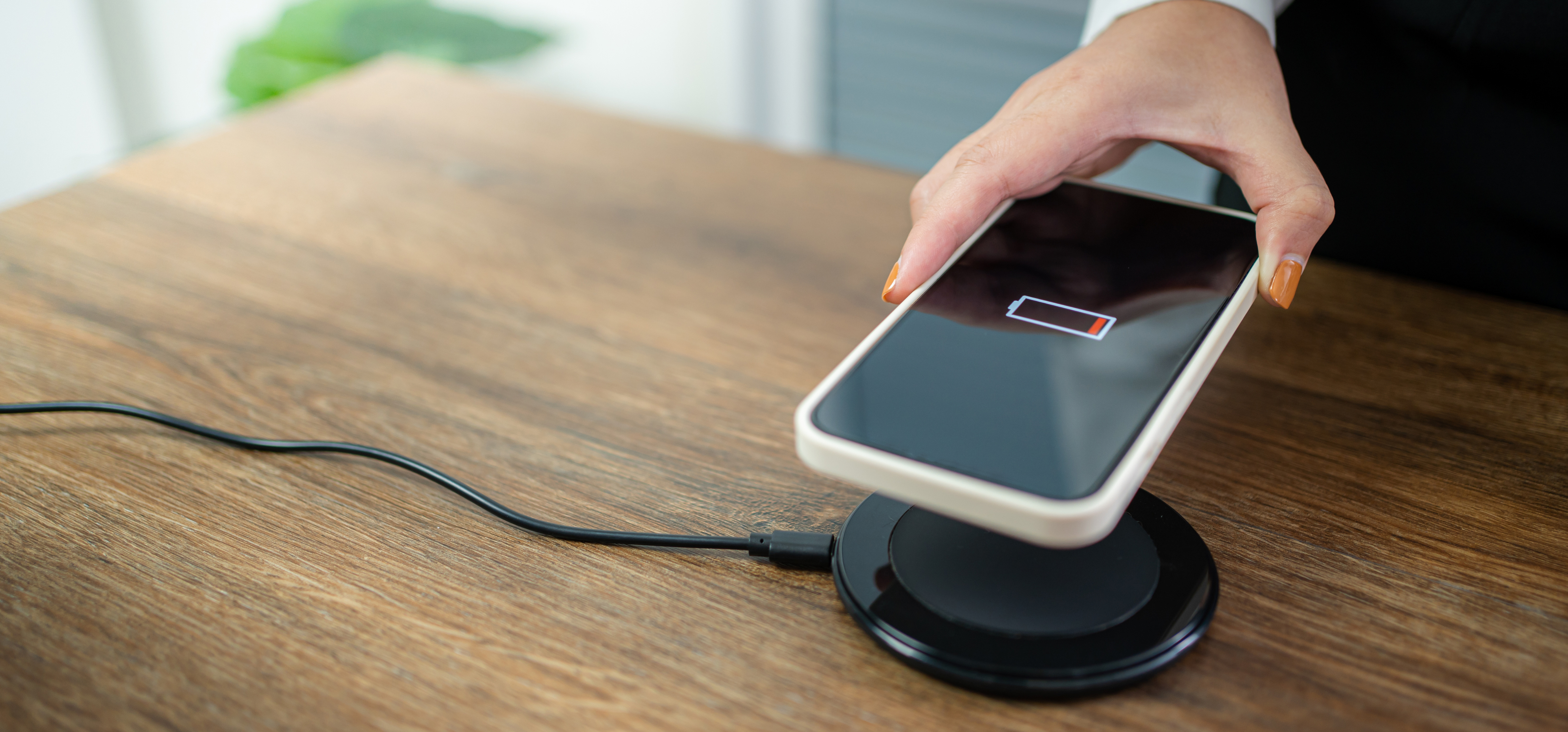 A woman with orange gel polish nail is charging her phone using NFC wireless charging on a wooden table.