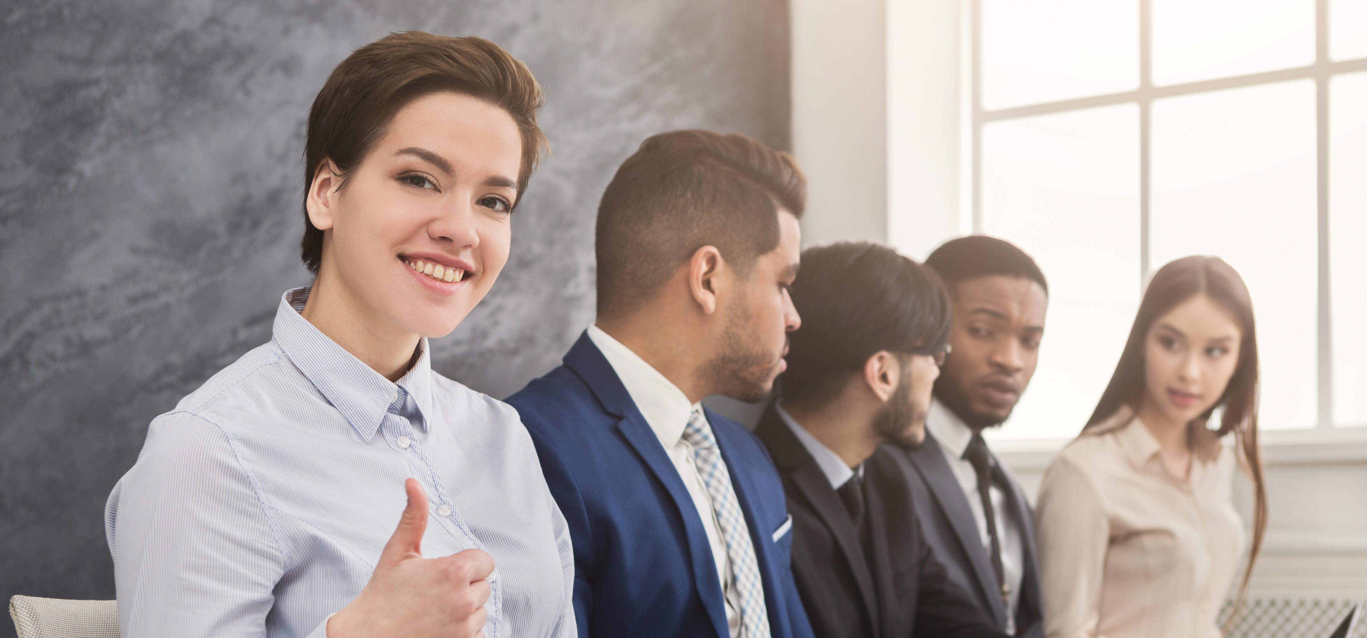 a woman with short hair wearing a light blue shirt gives a thumbs up to the camera and smile after successfully using ChatGPT to write a cover letter.