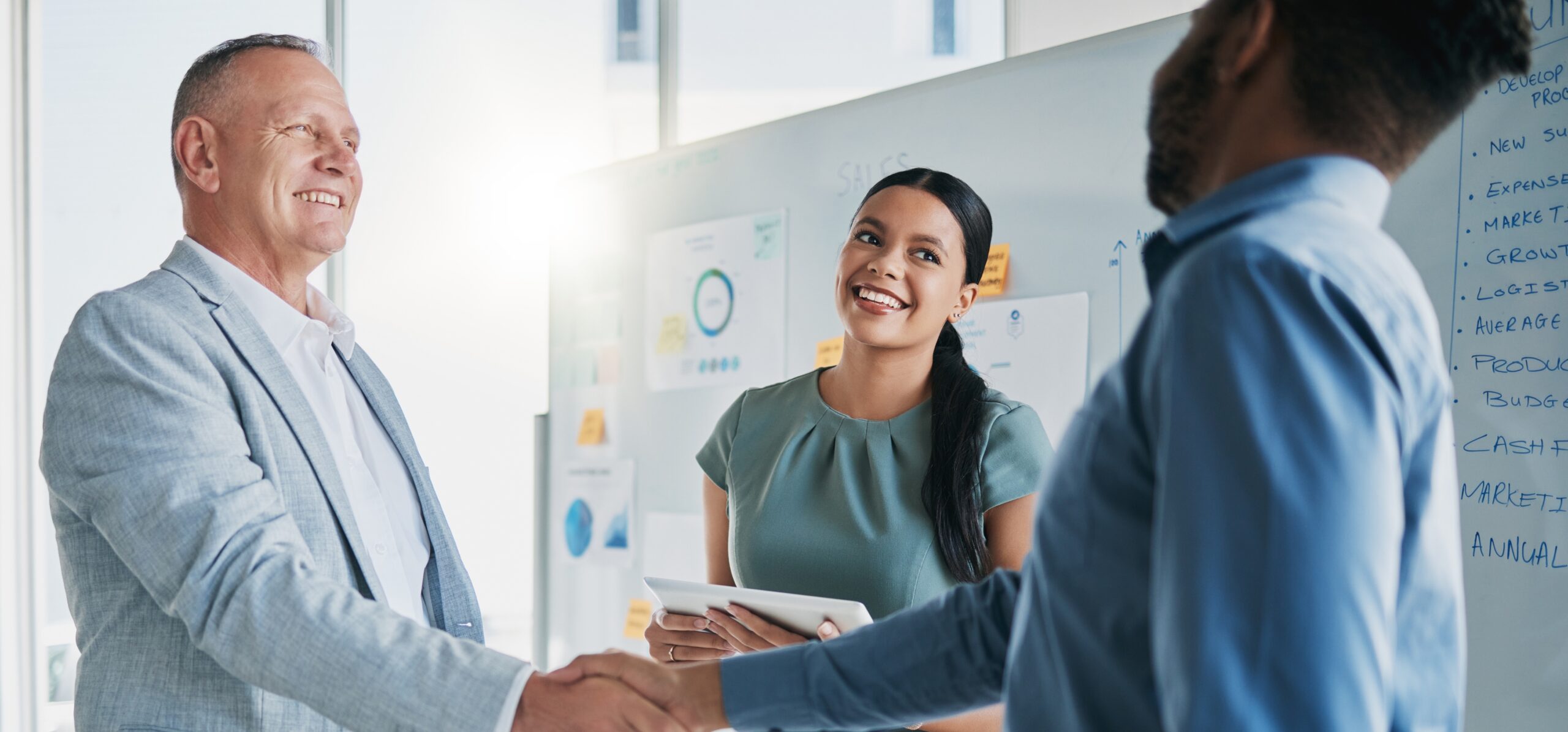 two directors from different companies are shaking hands signifying the business to business relationship marketing taking place between them while being witnessed by a female personal employee in a blue blouse.